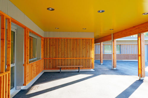 Extension et rénovation de l'école de Berville, par Atelier Cosme Architecture. Vue du préau, des façades bois et bac-acier, et de ses jeux d'ombres au sol.