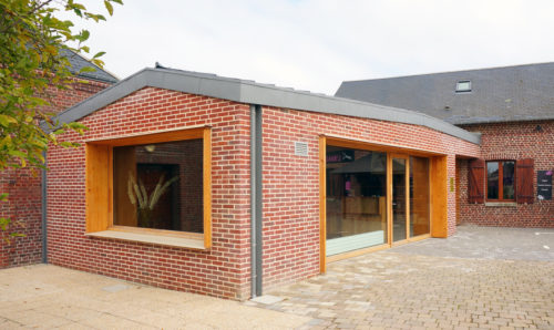 Construction d'un cabinet médical et d'un commerce par Atelier Cosme Architecture à Saint-Vigor-d'Ymonville. Vue d'ensemble des façades briques, menuiseries bois et toiture zinc.